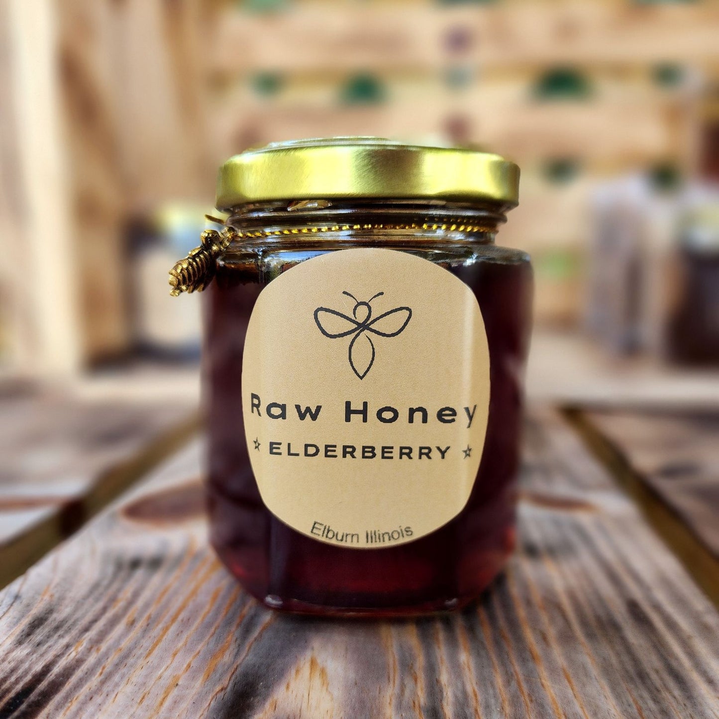 A glass jar filled with dark raw honey infused with elderberry, sitting on a rustic wooden table. The jar has a golden lid, and the label reads 'Raw Honey Elderberry,' with a simple bee icon above the text.