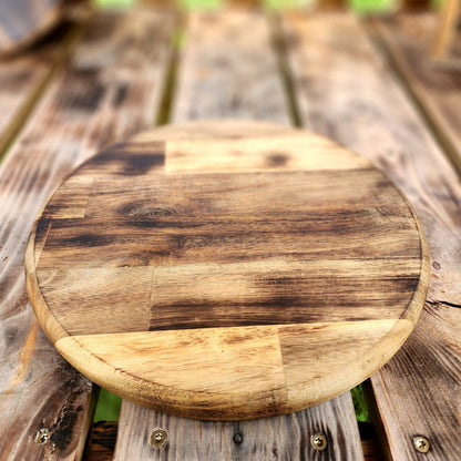 Round wooden cutting board with a natural, rustic finish featuring a blend of light and dark wood tones. The board sits on a weathered wooden table, showcasing a handcrafted and artisanal style, perfect for kitchen use or as a serving platter. Ideal for adding a touch of rustic charm to your kitchen décor.