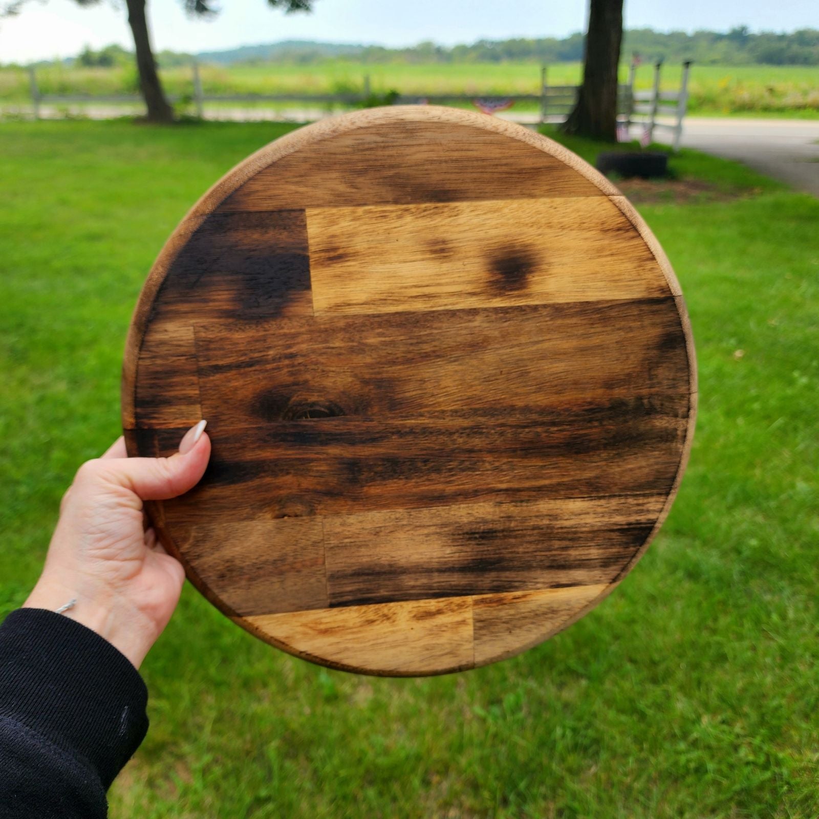 Hand holding a round wooden cutting board with a rustic finish. The board shows a mix of light and dark wood grains, highlighting its handcrafted, natural look. The background features a green, grassy field, giving the product an outdoor, farmhouse aesthetic. Ideal for kitchen use, serving, or décor.