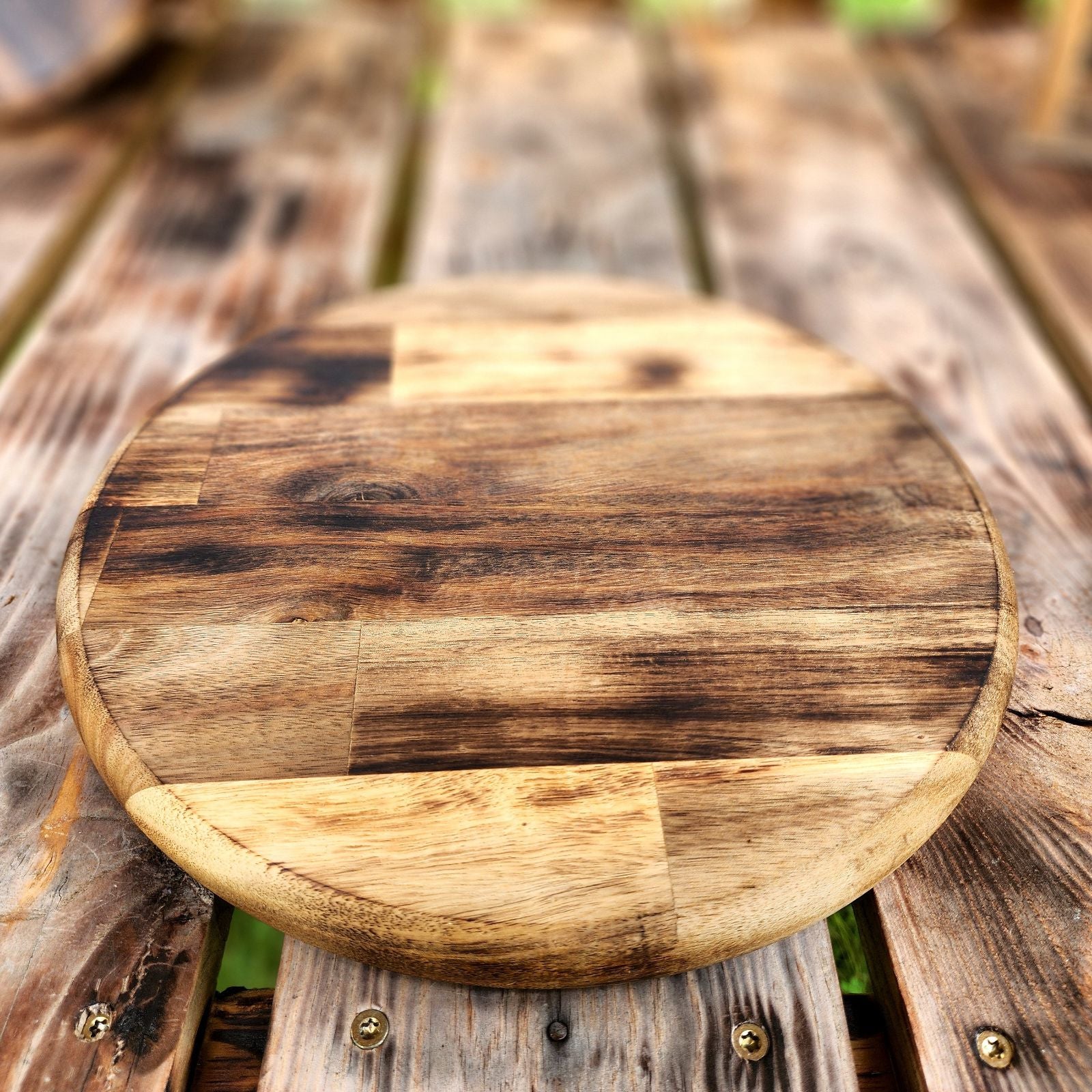 Round wooden cutting board with a rustic, natural finish featuring a blend of light and dark wood tones. The board is placed on a weathered wooden table, creating a farmhouse-style aesthetic. Ideal for serving or kitchen prep, this board adds charm and functionality to any kitchen.