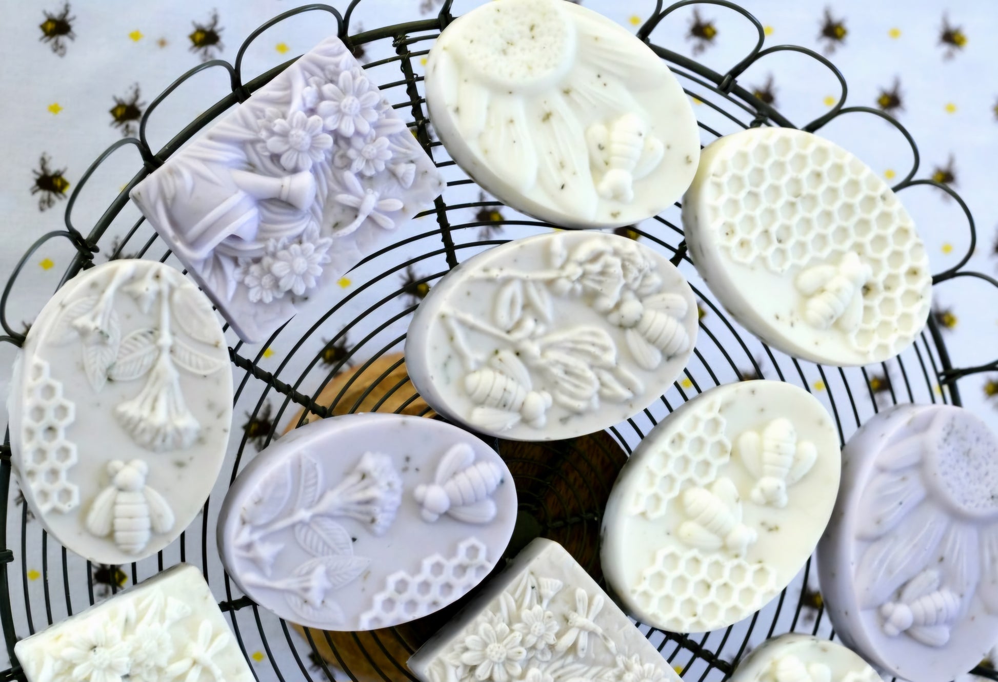 A display of handcrafted oval and rectangular soaps with detailed designs of bees, honeycomb, flowers, and leaves. The soaps are arranged on a circular wire rack, with a bee-patterned cloth in the background. The intricate carvings in the soaps highlight their artisanal quality and natural theme, making them perfect for eco-friendly and nature-inspired product collections.