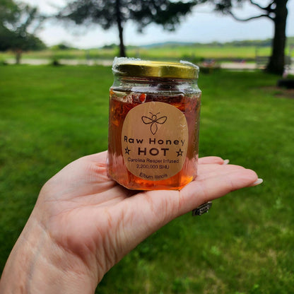 A hand holding a jar of Raw Honey Hot, infused with Carolina Reaper peppers, showcasing the honey’s label and vibrant outdoor setting in the background.