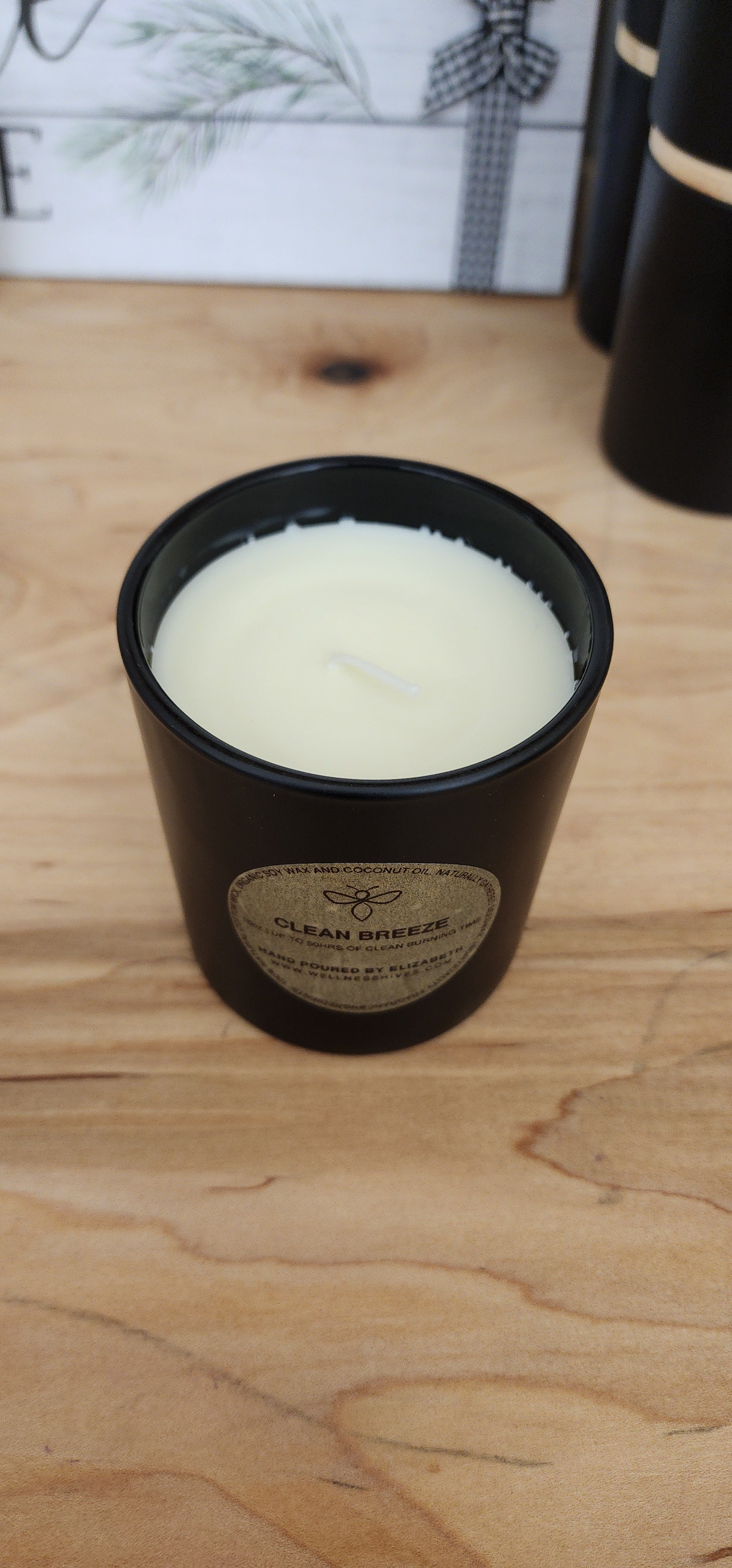 An open 'Clean Breeze' candle in a black jar with a gold label, filled with white wax. The candle, hand-poured by Elizabeth from wellnesshives.com, is made from organic soy wax and coconut oil, offering up to 50 hours of clean burning time