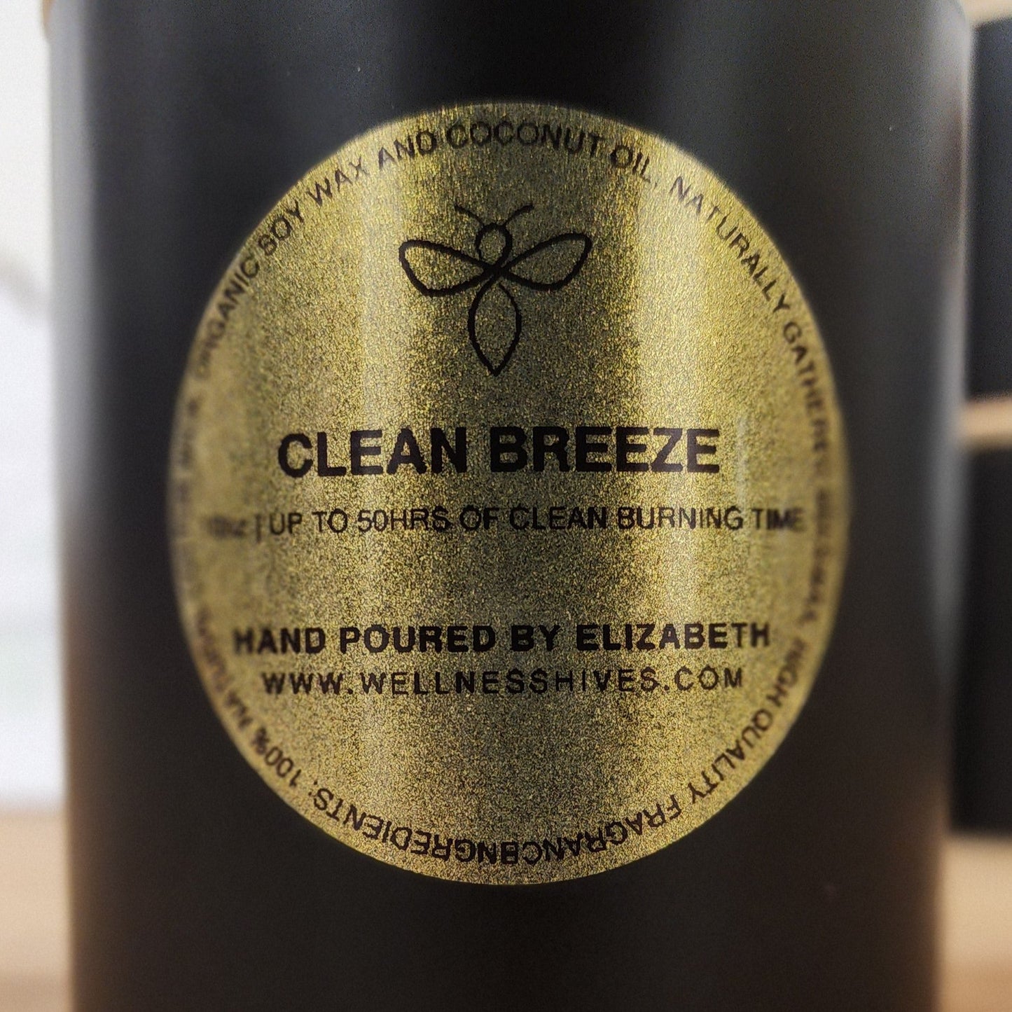 Close-up of the 'Clean Breeze' candle label on a black jar, highlighting 50 hours of clean burning time, hand-poured by Elizabeth from wellnesshives.com. The label emphasizes the use of organic soy wax and coconut oil with naturally gathered ingredients