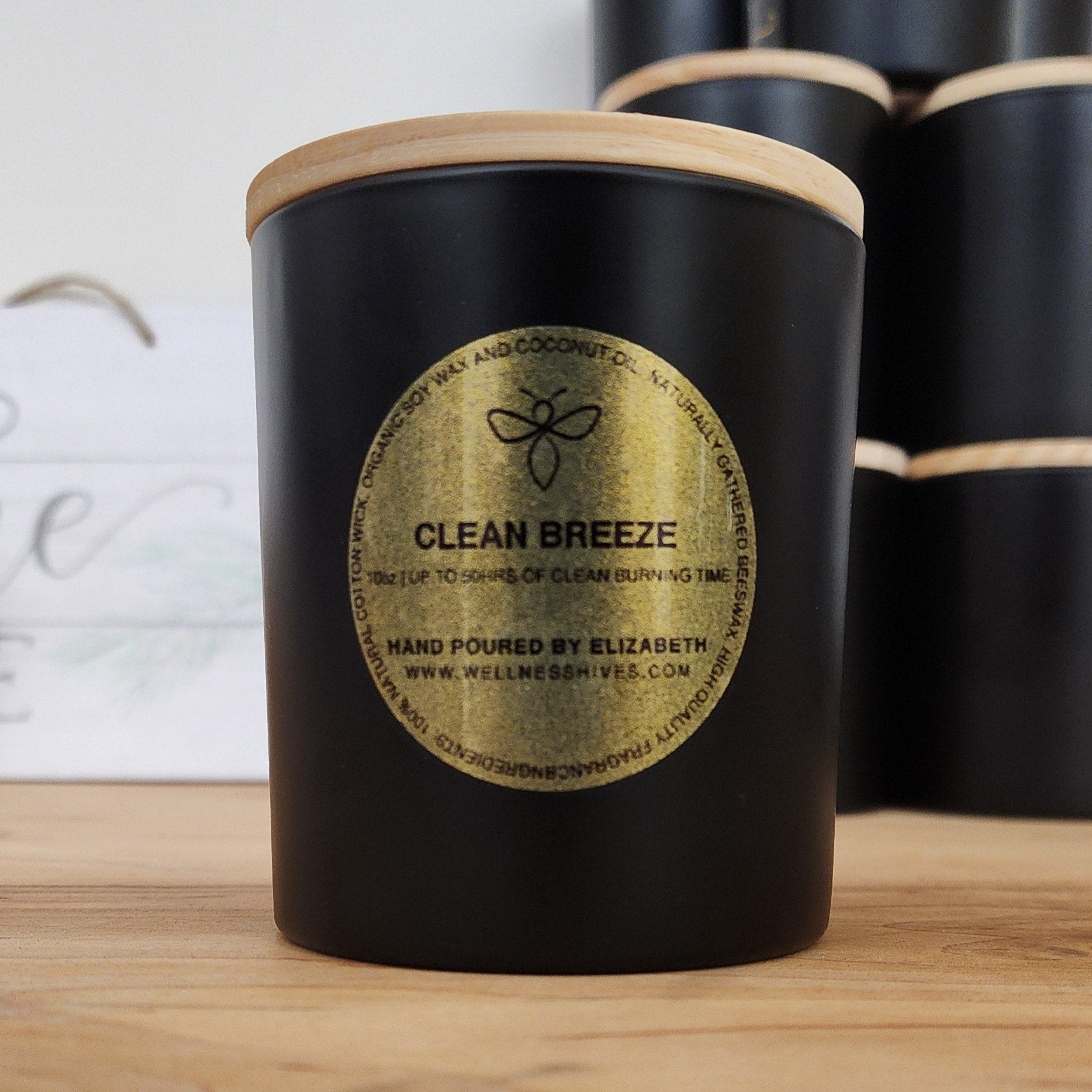 A black jar candle with a wooden lid, labeled 'Clean Breeze,' hand-poured by Elizabeth from wellnesshives.com. The label highlights its organic soy and coconut wax composition and clean burning time of up to 50 hours.