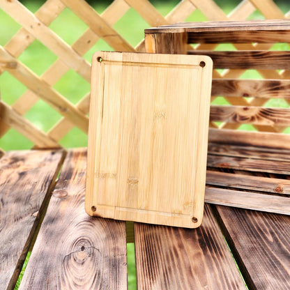 8" x 11" A rectangular bamboo cutting board displayed vertically on a rustic wooden table, with a lattice fence and a weathered wooden crate in the background. The cutting board features rounded corners with small holes in each corner for easy hanging or handling. The natural bamboo grain and clean design make it a functional and stylish addition to any kitchen.