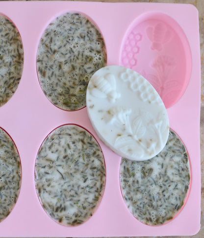 Oval-shaped artisan soaps in a silicone mold. Some of the soaps contain visible dried herbs, while others have intricate designs of bees, honeycomb, and flowers. The mix of textured and smooth soap surfaces showcases natural ingredients and handcrafted quality, perfect for promoting a rustic, eco-friendly product line.