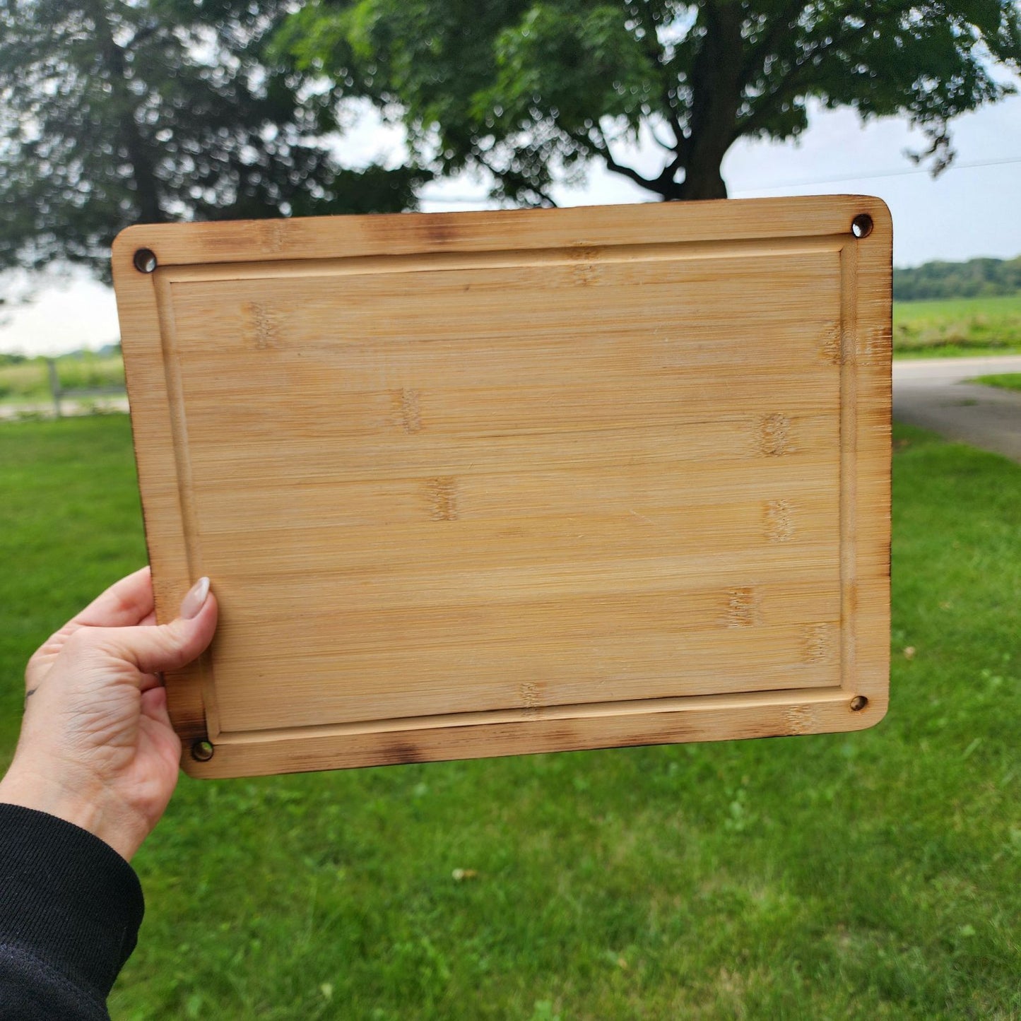 A hand holding a rectangular 9.5 x 13 bamboo cutting board with rounded corners and small holes in each corner. The cutting board is displayed outdoors against a backdrop of green grass and a scenic landscape. The natural bamboo grain and smooth finish emphasize its sturdy yet elegant design, perfect for kitchen tasks or serving food.
