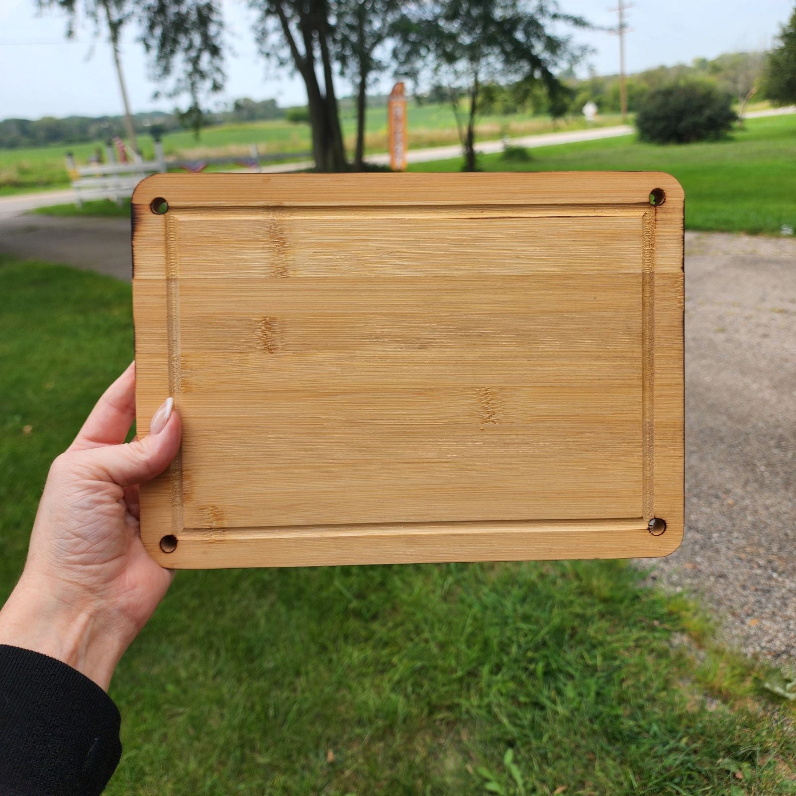 8" x 11" A hand holding a rectangular bamboo cutting board with rounded corners and small holes in each corner. The cutting board is displayed outdoors against a backdrop of green grass and a scenic landscape. The natural bamboo grain and smooth finish emphasize its sturdy yet elegant design, perfect for kitchen tasks or serving food.