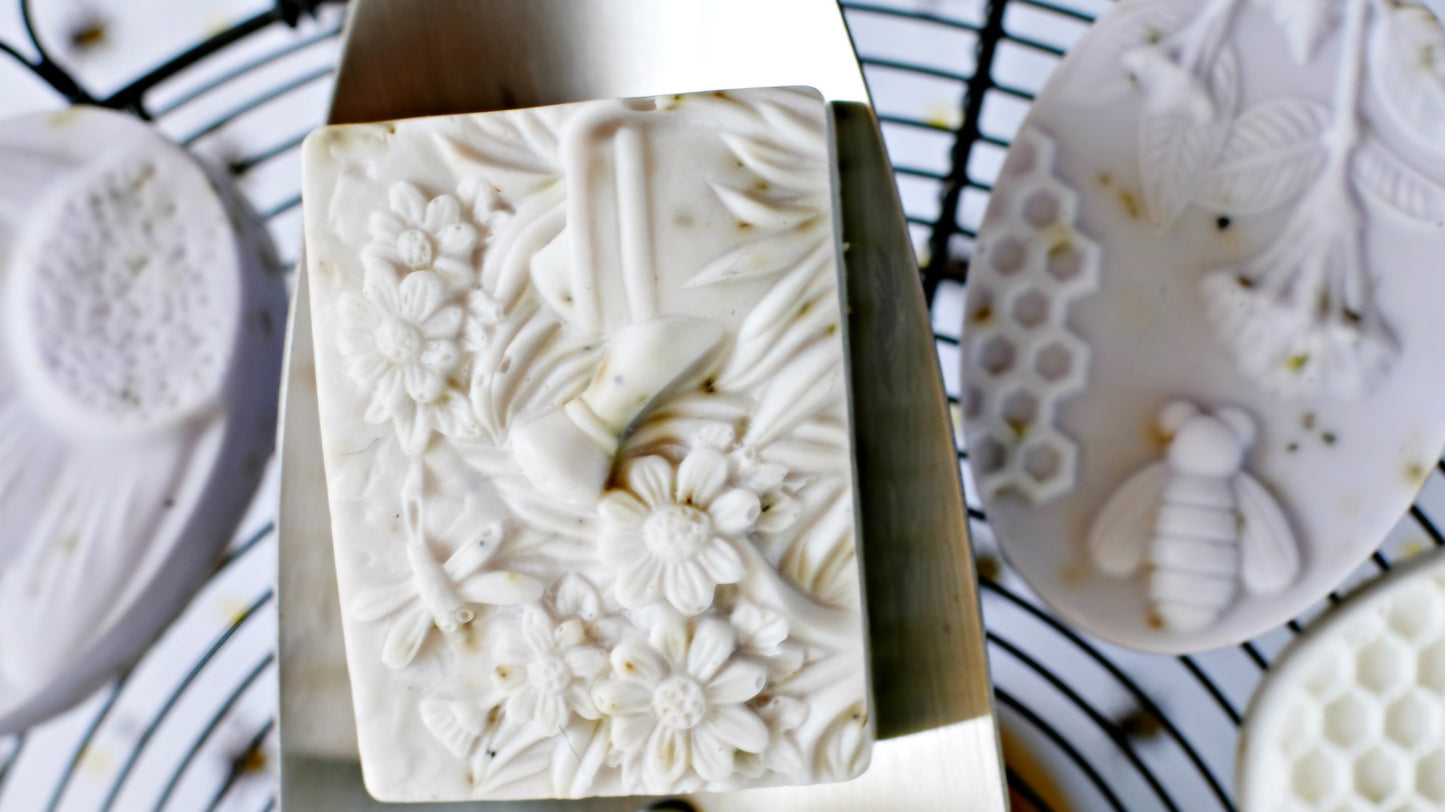 A close-up of a rectangular handcrafted soap featuring an intricate floral and dragonfly design. The soap is surrounded by other oval-shaped soaps with bee and honeycomb patterns, displayed on a wire rack. The detailed carvings emphasize the artisanal craftsmanship and natural inspiration behind the soap collection, ideal for eco-conscious buyers.
