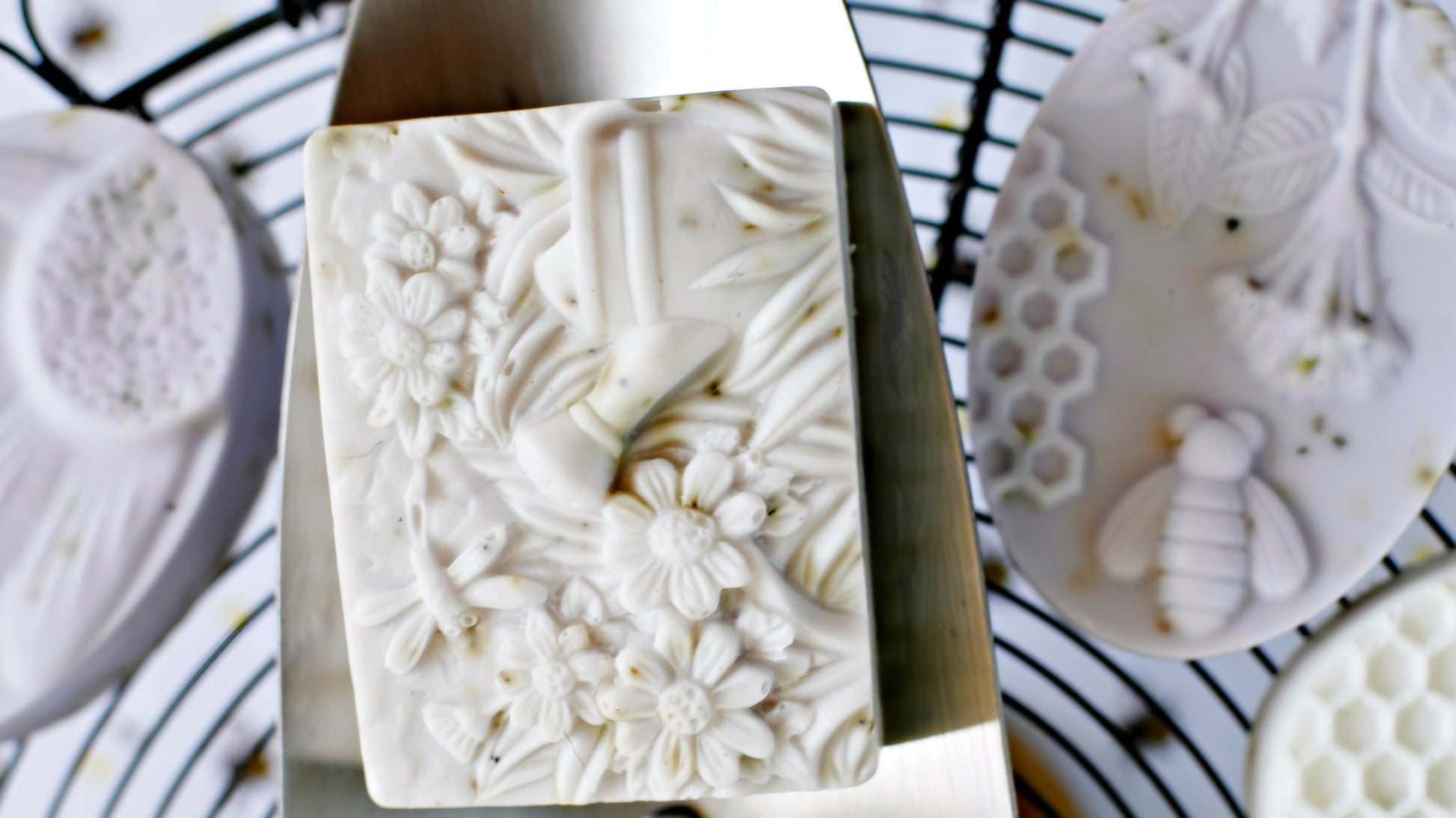 A close-up of a rectangular handcrafted soap featuring an intricate floral and dragonfly design. The soap is surrounded by other oval-shaped soaps with bee and honeycomb patterns, displayed on a wire rack. The detailed carvings emphasize the artisanal craftsmanship and natural inspiration behind the soap collection, ideal for eco-conscious buyers.
