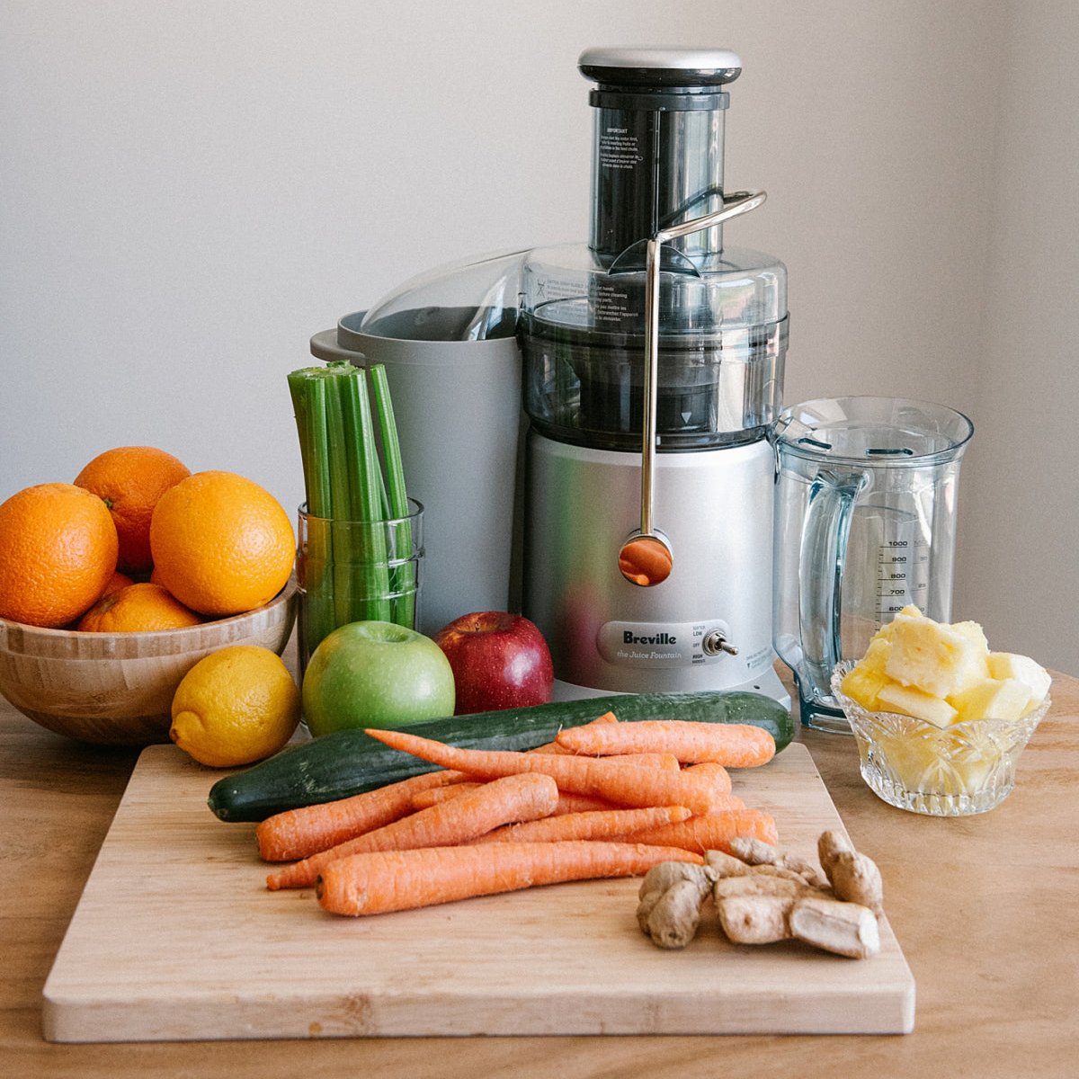 A variety of fresh fruits and vegetables, including carrots, ginger, oranges, celery, cucumber, and pineapple, displayed on a kitchen counter next to a juicer, ready for juicing as part of a health and wellness routine