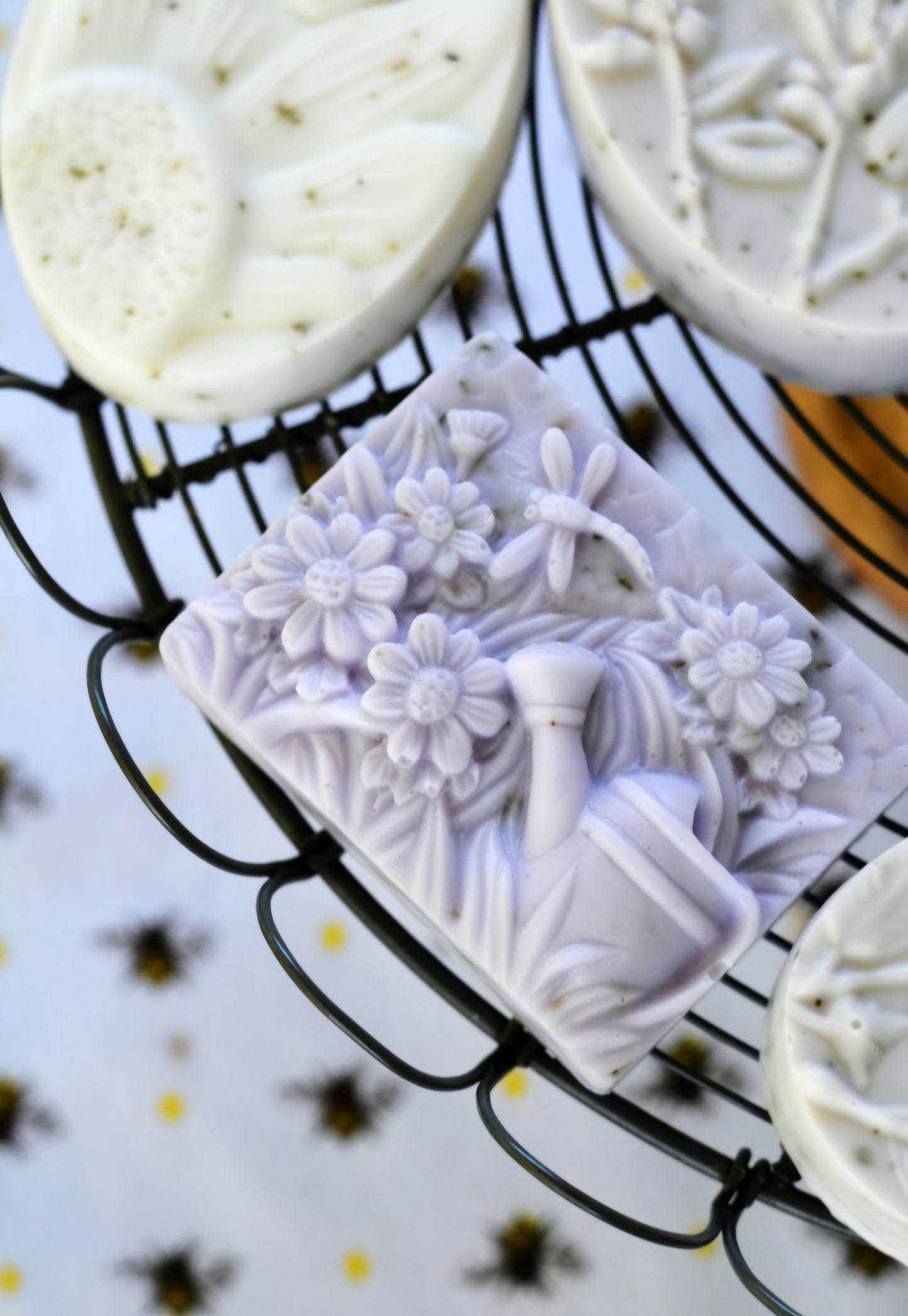 A close-up of beautifully detailed rectangular artisan soap featuring a floral design with daisies and leaves. The soap is placed on a wire rack alongside other oval-shaped soaps with intricate patterns. The background showcases a bee-themed cloth, enhancing the natural, eco-friendly appeal of the handcrafted soaps.