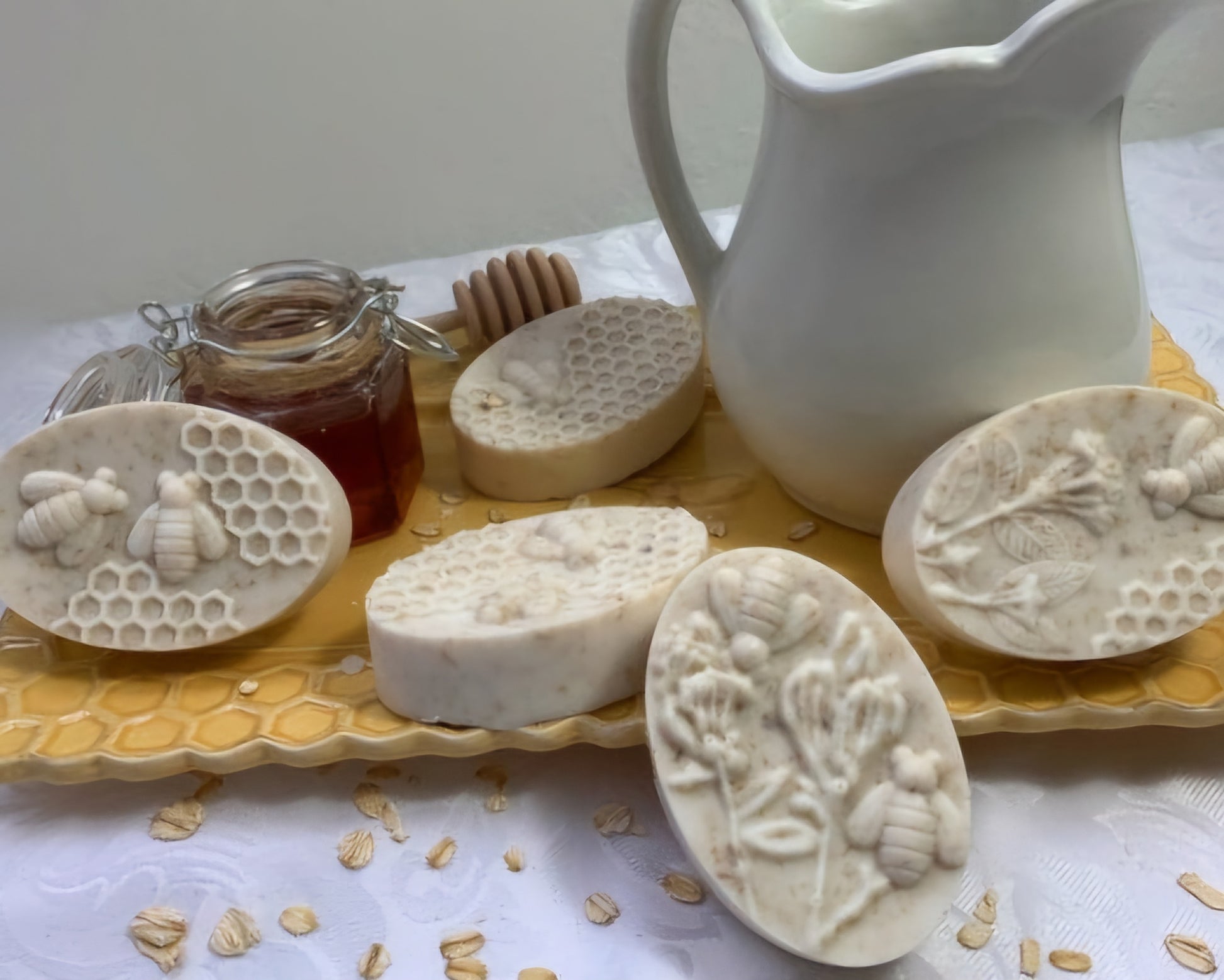 A beautifully styled display of bee and honeycomb-themed artisan soaps on a honeycomb-patterned tray. Accompanying the soaps are a jar of honey with a wooden dipper and a white ceramic pitcher, adding to the rustic, nature-inspired scene. The soaps feature intricate designs of bees, honeycomb, and flowers, with oats scattered across the table, evoking a sense of natural ingredients and handcrafted luxury.