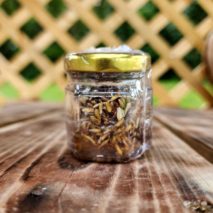 A glass jar filled with honey infused with organic lavender, sealed with a golden lid, sitting on a wooden surface against a blurred lattice background.