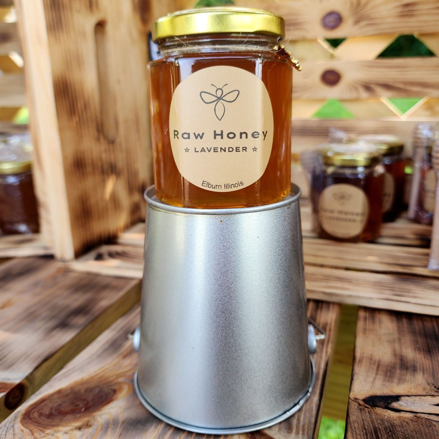 A 10 oz jar of Raw Lavender Honey, placed on a metallic stand, is displayed in an indoor wooden setting. The label clearly reads 'Raw Honey Lavender,' and the honey inside glows a rich amber color. Other honey jars can be seen blurred in the background, adding a rustic and warm atmosphere to the display