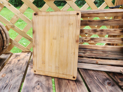 A rectangular 9.5 x 13 bamboo cutting board displayed outdoors on a rustic wooden table, with a lattice fence and wooden crate in the background. The cutting board features rounded corners with small holes, adding a touch of design to its simple and functional look. The natural bamboo grain and smooth finish highlight its handcrafted quality, ideal for kitchen use or as a serving board.
