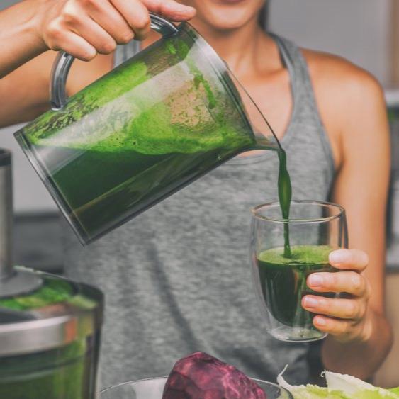 A woman pouring fresh green juice from a glass pitcher into a glass, preparing a nutritious drink. The scene emphasizes healthy living and juicing as part of a wellness routine.